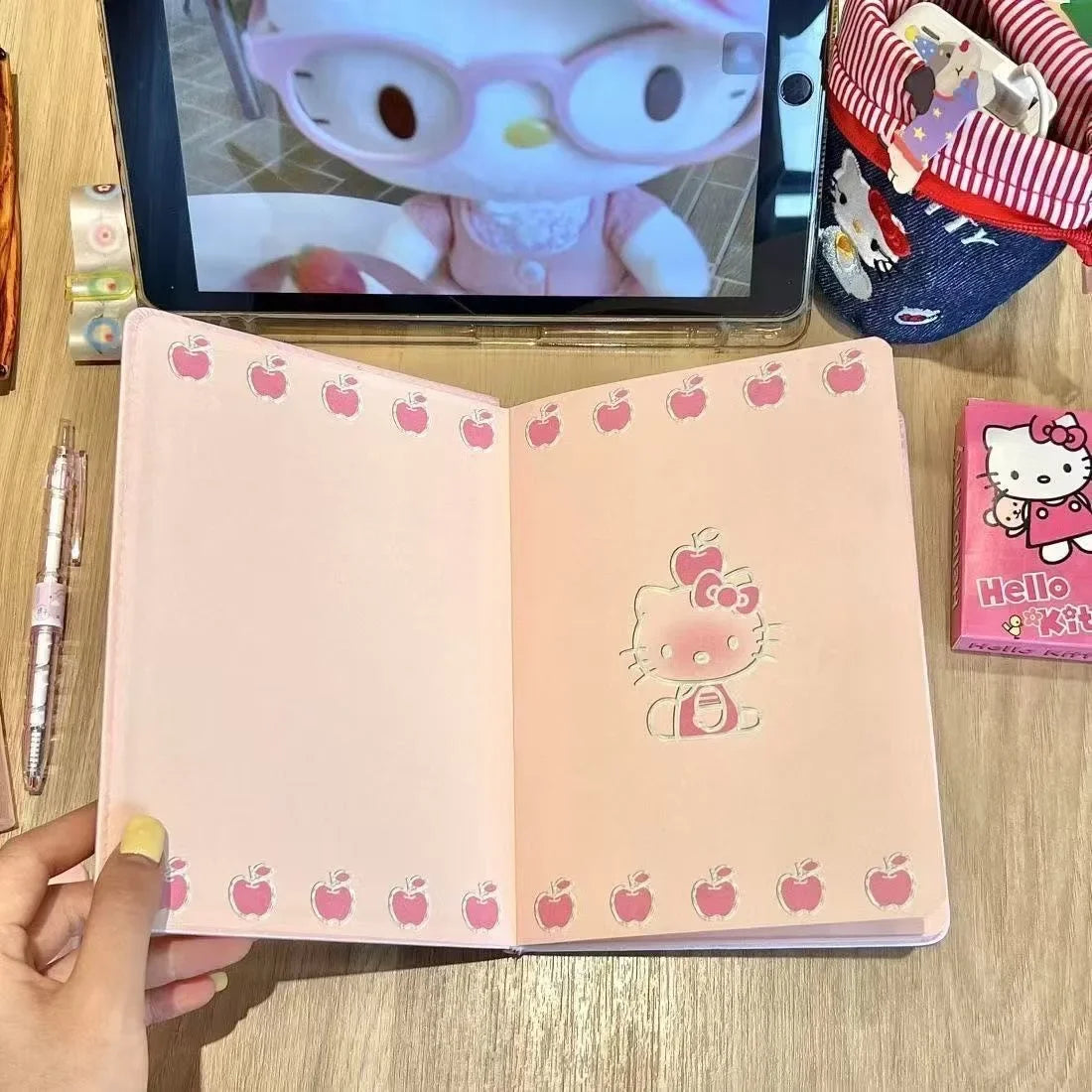 Notebook with Hello Kitty design on a wooden surface, with a tablet displaying a cartoon character in the background.
