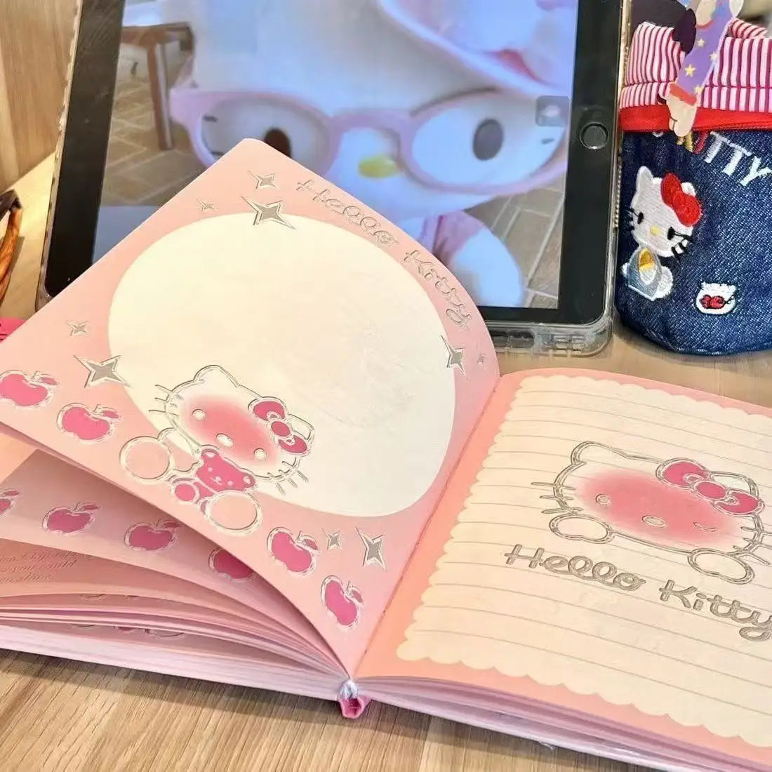 Hello Kitty notebook open on a wooden surface with a tablet displaying a cartoon character in the background.