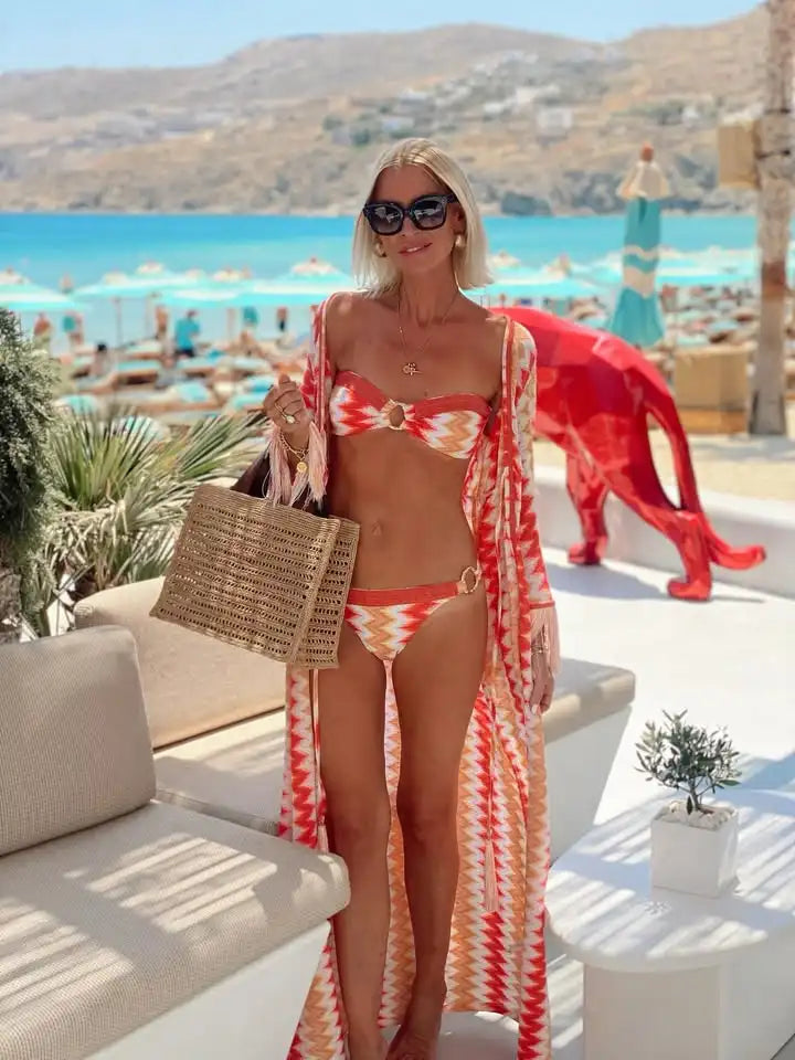 Woman in a colorful bikini and cover-up standing on a sunlit patio with a beach in the background.