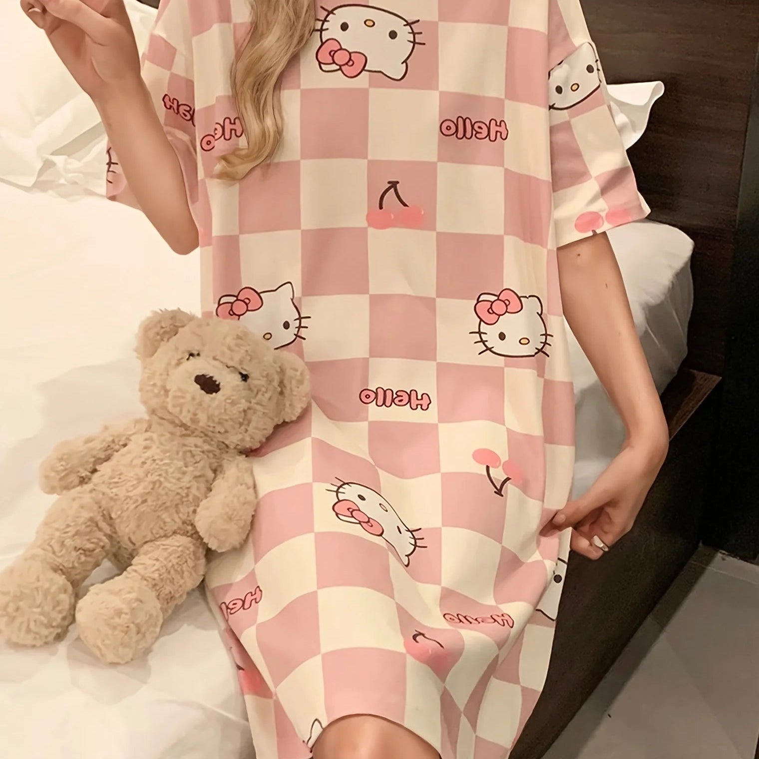Person wearing a pink checkered nightgown with cartoon characters, sitting on a bed with a teddy bear.