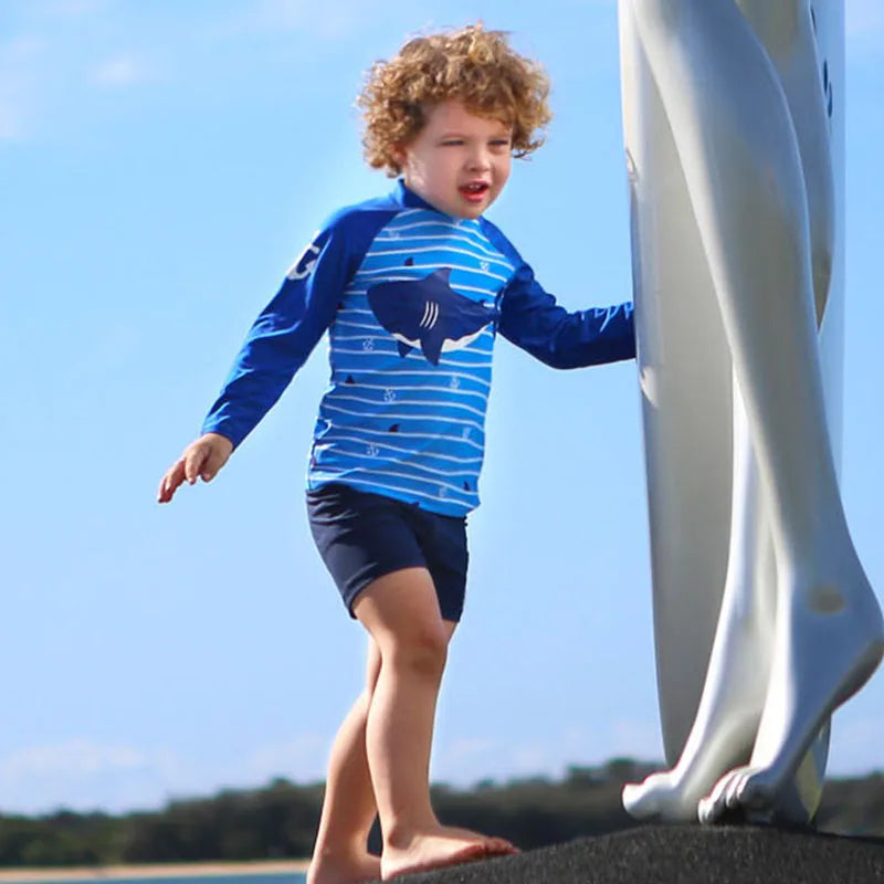 Child wearing a blue shark-themed swimsuit standing near a sculpture with a clear sky.