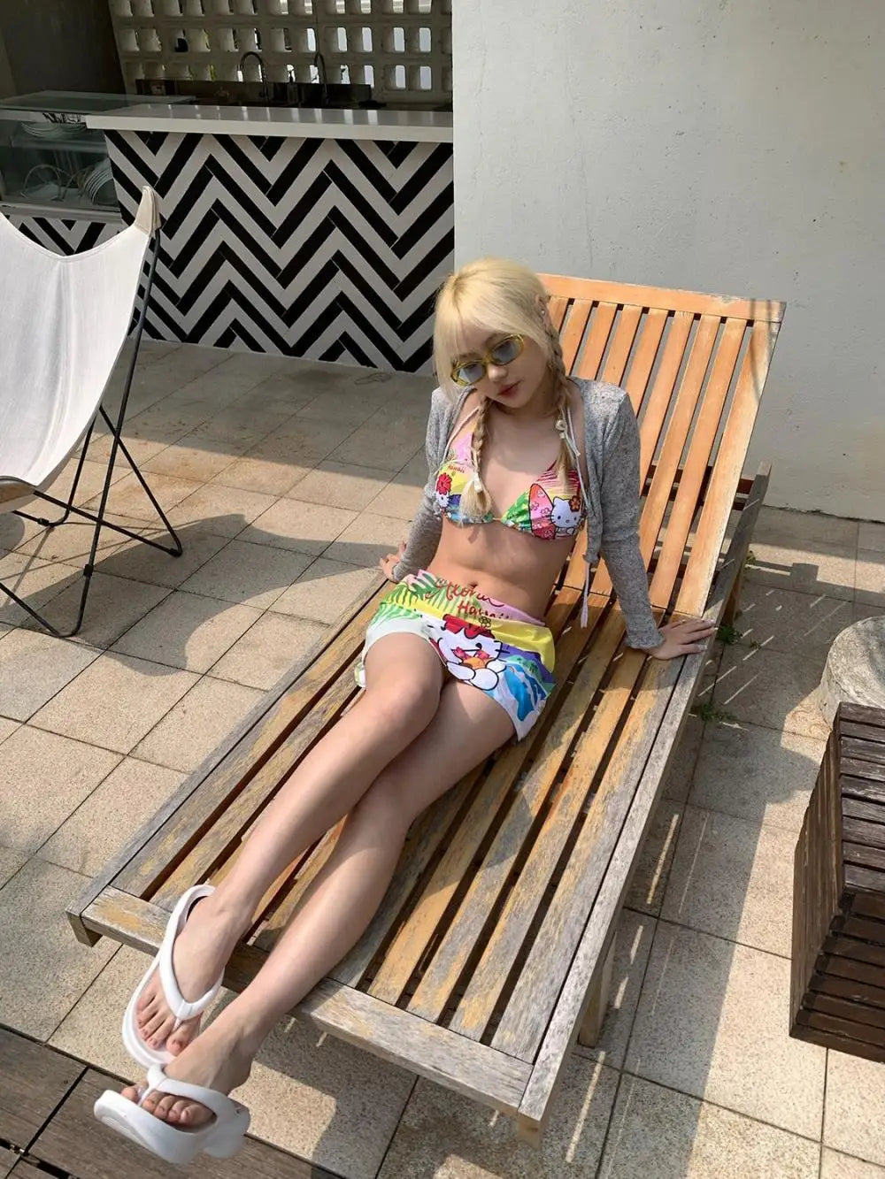 Woman in a colorful bikini and gray cardigan lying on a wooden bench outdoors.