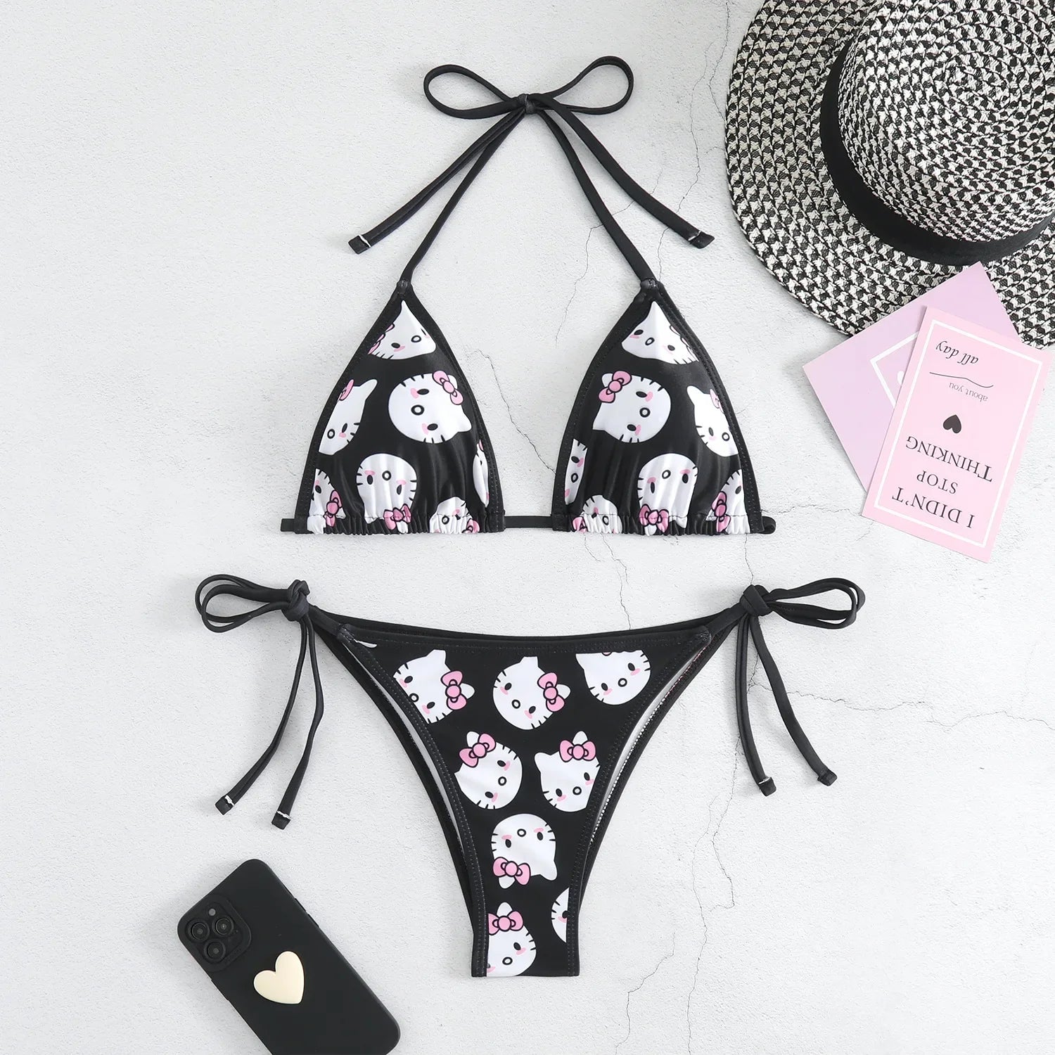Black bikini with white cat print on a white surface with a hat and phone case.
