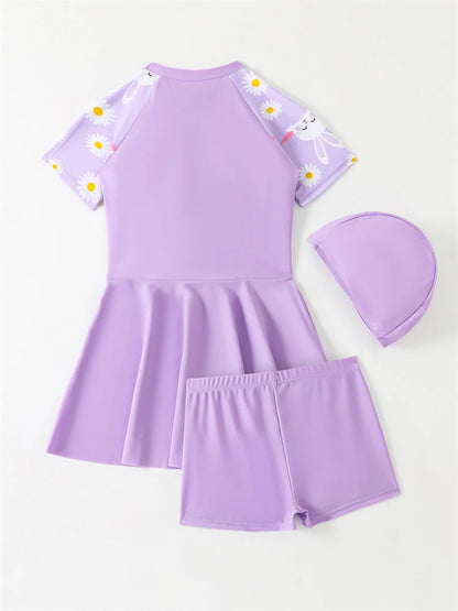 Purple children's swimsuit set with a dress, shorts, and cap on a white background