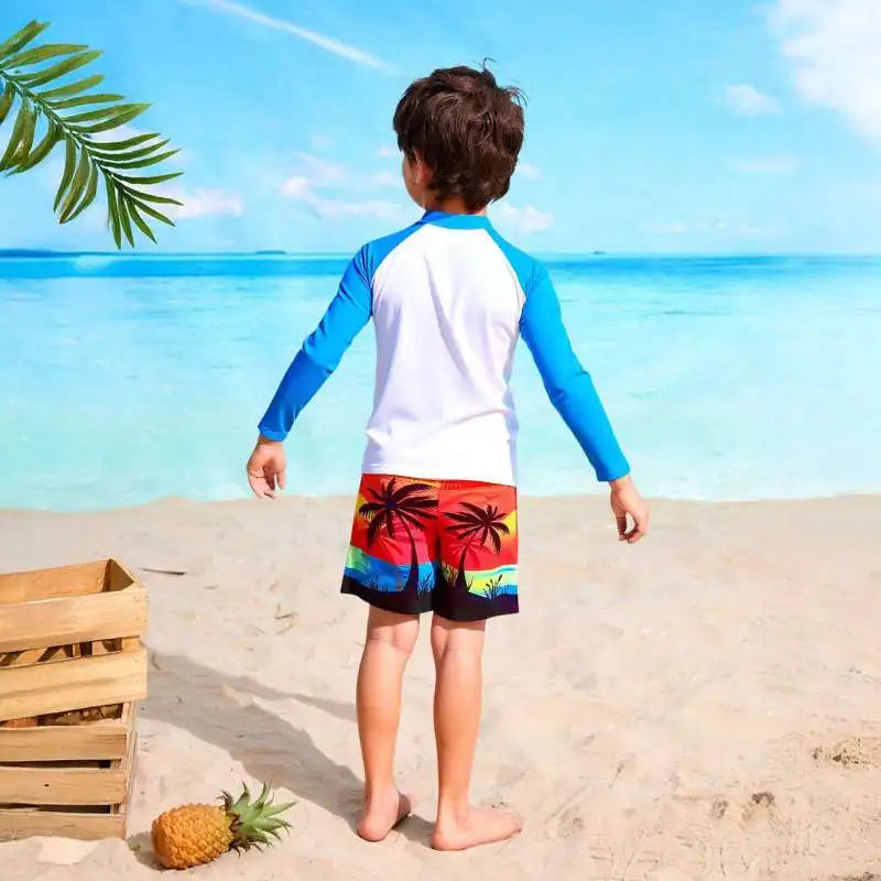 Child wearing a blue and white long-sleeve shirt and colorful shorts standing on a sandy beach with ocean view.